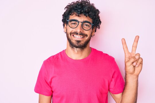 Handsome young man with curly hair and bear wearing casual clothes and glasses smiling with happy face winking at the camera doing victory sign. number two.