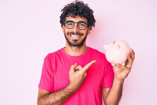 Handsome young man with curly hair and bear holding piggy bank smiling happy pointing with hand and finger