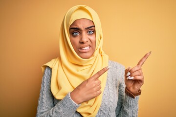 Young beautiful african american girl wearing muslim hijab over isolated yellow background Pointing aside worried and nervous with both hands, concerned and surprised expression