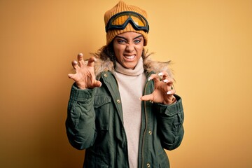 Young african american afro skier girl wearing snow sportswear and ski goggles smiling funny doing claw gesture as cat, aggressive and sexy expression