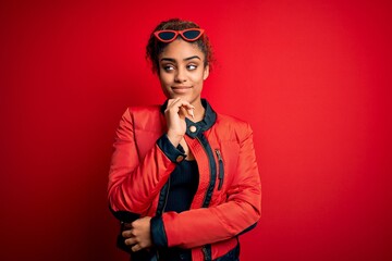 Beautiful african american girl wearing red jacket and sunglasses over isolated background with hand on chin thinking about question, pensive expression. Smiling with thoughtful face. Doubt concept.