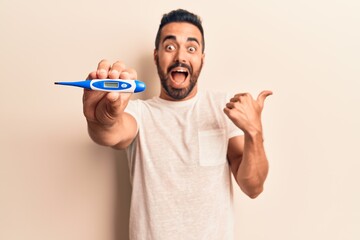 Young hispanic man holding thermometer pointing thumb up to the side smiling happy with open mouth