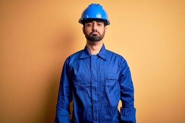 Mechanic man with beard wearing blue uniform and safety helmet over yellow background depressed and worry for distress, crying angry and afraid. Sad expression.