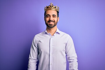 Young handsome man with beard wearing golden crown of king over purple background with a happy and cool smile on face. Lucky person.