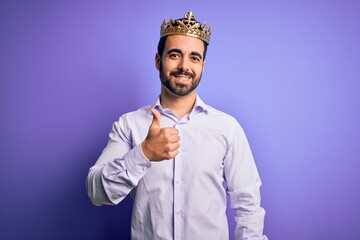 Young handsome man with beard wearing golden crown of king over purple background doing happy thumbs up gesture with hand. Approving expression looking at the camera showing success.