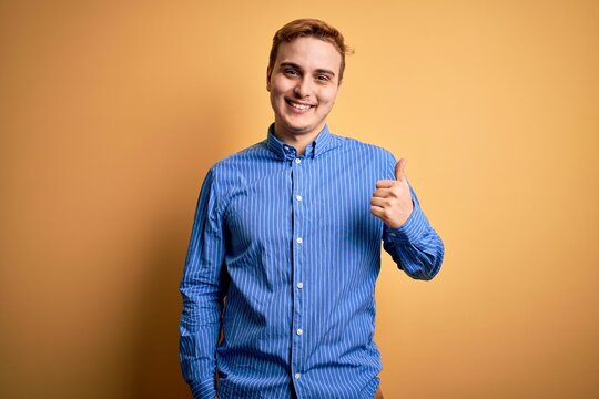 Young handsome redhead man wearing casual striped shirt over isolated yellow background doing happy thumbs up gesture with hand. Approving expression looking at the camera showing success.