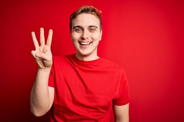 Young handsome redhead man wearing casual t-shirt over isolated red background showing and pointing up with fingers number three while smiling confident and happy.