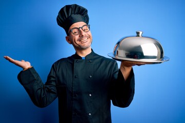 Young handsome chef man wearing uniform and hat holding waiter tray with dome very happy and excited, winner expression celebrating victory screaming with big smile and raised hands
