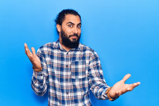 Young arab man wearing casual clothes clueless and confused expression with arms and hands raised. doubt concept.