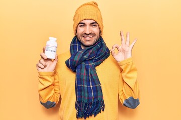 Young handsome bald man wearing winter clothes holding jar of pills doing ok sign with fingers, smiling friendly gesturing excellent symbol