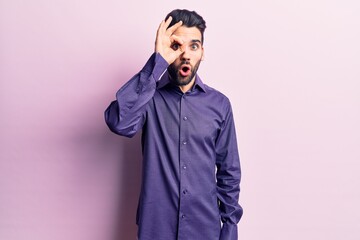 Young handsome man with beard wearing casual shirt doing ok gesture shocked with surprised face, eye looking through fingers. unbelieving expression.