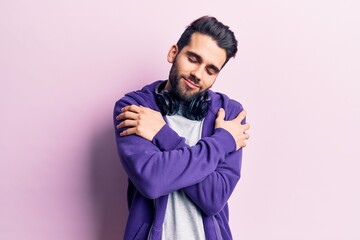 Young handsome man with beard listening to music using headphones hugging oneself happy and...