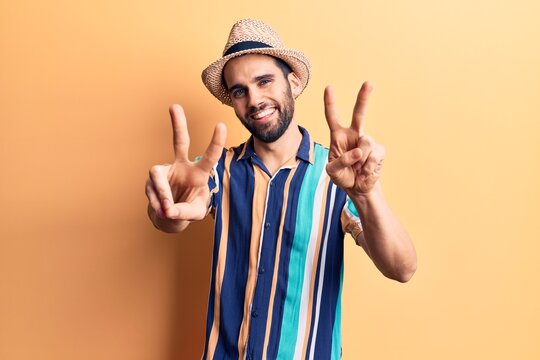 Young handsome man with beard wearing summer hat and shirt smiling looking to the camera showing fingers doing victory sign. number two.