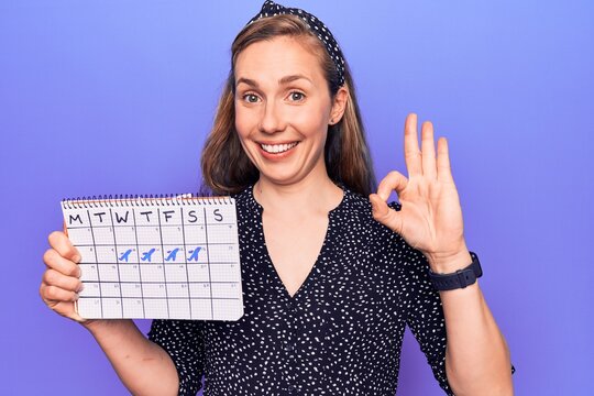 Young beautiful blonde woman holding holidays trip calendar doing ok sign with fingers, smiling friendly gesturing excellent symbol