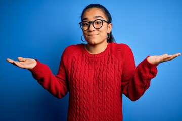 Young beautiful asian woman wearing casual sweater and glasses over blue background clueless and...