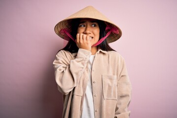Fototapeta premium Young beautiful woman wearing traditional conical asian hat over isolated pink background looking stressed and nervous with hands on mouth biting nails. Anxiety problem.