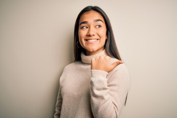 Young beautiful asian woman wearing casual turtleneck sweater over white background smiling with happy face looking and pointing to the side with thumb up.