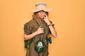 Young blonde explorer woman with blue eyes hiking wearing backpack and water canteen Pointing to the eye watching you gesture, suspicious expression