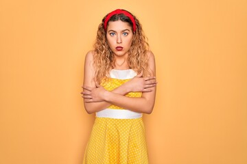 Beautiful blonde pin-up woman with blue eyes wearing diadem standing over yellow background shaking and freezing for winter cold with sad and shock expression on face