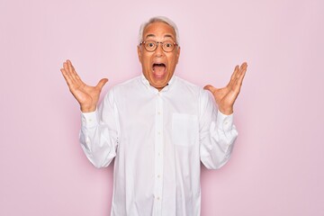 Middle age senior grey-haired man wearing glasses and business shirt over pink background celebrating crazy and amazed for success with arms raised and open eyes screaming excited. Winner concept