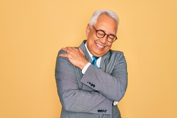 Middle age senior grey-haired handsome business man wearing glasses over yellow background Hugging oneself happy and positive, smiling confident. Self love and self care