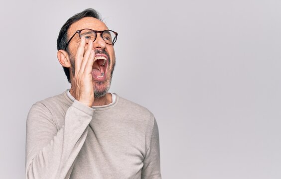 Middle age handsome man wearing casual sweater and glasses over isolated white background shouting and screaming loud to side with hand on mouth. Communication concept.