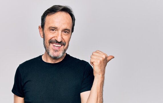 Middle age man wearing casual black t-shirt standing over isolated white background pointing thumb up to the side smiling happy with open mouth
