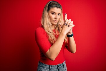Young beautiful blonde woman wearing casual t-shirt standing over isolated red background Holding symbolic gun with hand gesture, playing killing shooting weapons, angry face