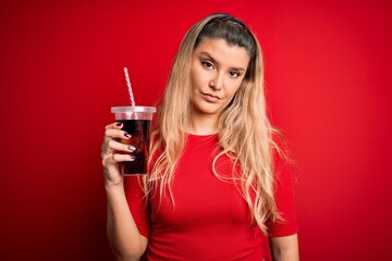 Young beautiful blonde woman drinking cola fizzy beverage using straw over red background with a confident expression on smart face thinking serious