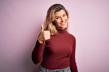 Young beautiful blonde woman wearing casual sweater over isolated pink background doing happy thumbs up gesture with hand. Approving expression looking at the camera showing success.