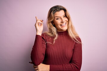 Fototapeta na wymiar Young beautiful blonde woman wearing casual sweater over isolated pink background with a big smile on face, pointing with hand and finger to the side looking at the camera.