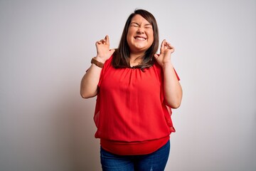 Beautiful brunette plus size woman wearing casual red t-shirt over isolated white background gesturing finger crossed smiling with hope and eyes closed. Luck and superstitious concept.