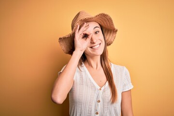 Young beautiful woman wearing casual t-shirt and summer hat over isolated yellow background doing ok gesture with hand smiling, eye looking through fingers with happy face.