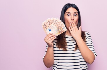 Young beautiful brunette woman holding euros banknotes over isolated pink background covering mouth...