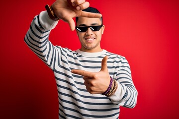 Naklejka premium Young brazilian man wearing funny thug life sunglasses over isolated red background smiling making frame with hands and fingers with happy face. Creativity and photography concept.
