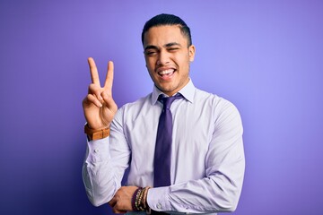 Young brazilian businessman wearing elegant tie standing over isolated purple background smiling with happy face winking at the camera doing victory sign with fingers. Number two.