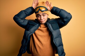 Young brazilian skier man wearing snow sportswear and ski goggles over yellow background Doing...