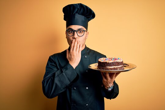 Young brazilian baker man wearing cooker uniform and hat holding tray with cake cover mouth with hand shocked with shame for mistake, expression of fear, scared in silence, secret concept