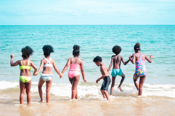 Group of six boys and girls standing in a line in shallow sea water, cute kids having fun on sandy summer beach, happy childhood friend playing on tropical sea