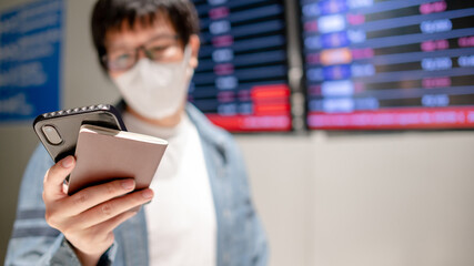 Asian man tourist wearing face mask holding smartphone and passport at arrival departure board in airport terminal. Coronavirus (COVID-19) pandemic prevention when travel. Social distancing concept