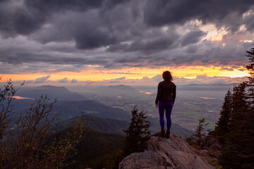 Fototapeta premium Adventurous Girl on top of a Rocky Mountain overlooking the beautiful Canadian Nature Landscape during a dramatic Sunset. Taken in Chilliwack, East of Vancouver, British Columbia, Canada.