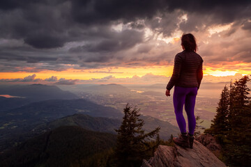 Obraz premium Adventurous Girl on top of a Rocky Mountain overlooking the beautiful Canadian Nature Landscape during a dramatic Sunset. Taken in Chilliwack, East of Vancouver, British Columbia, Canada.
