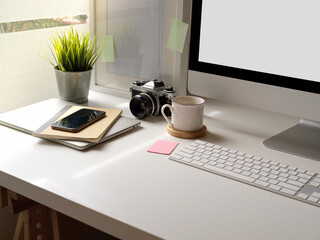 Fototapeta premium Modern office desk with mock-up computer, smartphone, camera and office supplies beside window