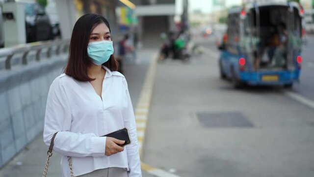 young asian woman holding smartphone and waiting for bus at bus stop in city street and wearing face mask protective for spreading of coronavirus(covid-19) pandemic, new normal concept