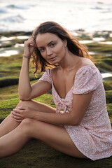 Beautiful young girl in a dress sits on stones on the ocean at sunset. A full-length girl is looking at the camera. Portrait of a brown-haired young woman.