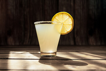 summer alcoholic yellow frosted drink in a transparent shaped glass with sugar on the rim and a round ripe juicy slice of lemon on a wooden table, photo for the bar menu