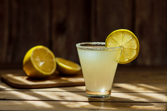 summer alcoholic yellow frosted drink in a transparent shaped glass with sugar on the rim and a round ripe juicy slice of lemon on a wooden table and round lemons in the background