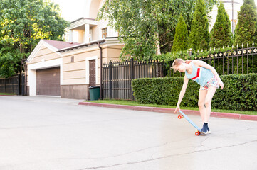 Teen girl rides on a skateboard in the street in the fresh air. Clothing: blue T-shirt, white shorts with a flower pattern. The board is blue with red wheels. on the street asphalt, trees, home