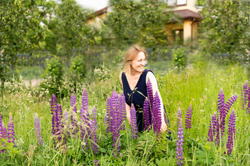 A girl with blond hair is standing in tall grass, looking to the side, laughing and beautiful blooming purple lupins, lilac shades in the field, a woman has a long blue silk dress, brown eyes