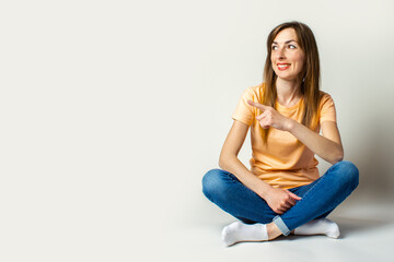Young woman in casual clothes looks away and points a finger while sitting on the floor on a light background